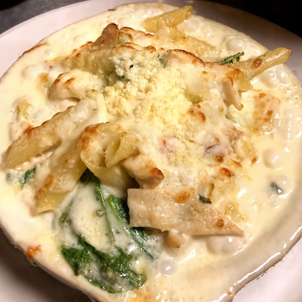 Baked penne pasta with creamy white sauce, melted cheese, and spinach in a white dish.
