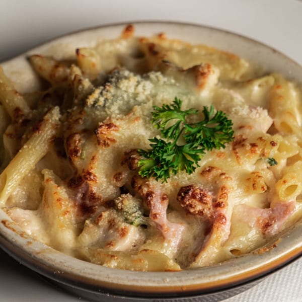 Baked penne pasta with melted cheese, breadcrumbs, and parsley in a ceramic dish.