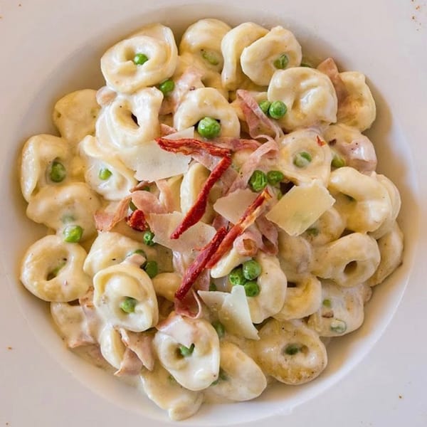A white bowl filled with creamy tortellini pasta, peas, bacon, and parmesan cheese.