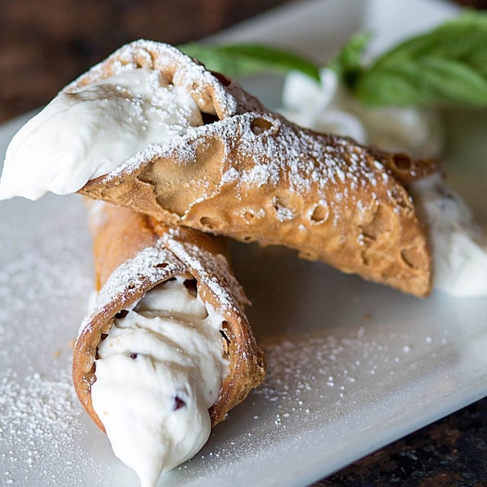 Two cannolis filled with white cream and chocolate chips, dusted with powdered sugar on a white plate.